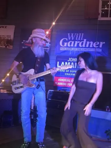 Dino performing guitar at a neon-lit venue with a fan dancing